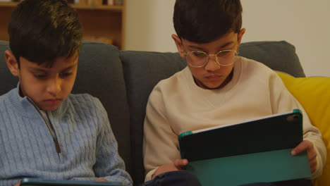 Two-Young-Boys-Sitting-On-Sofa-At-Home-Playing-Games-Or-Streaming-Onto-Digital-Tablets-4